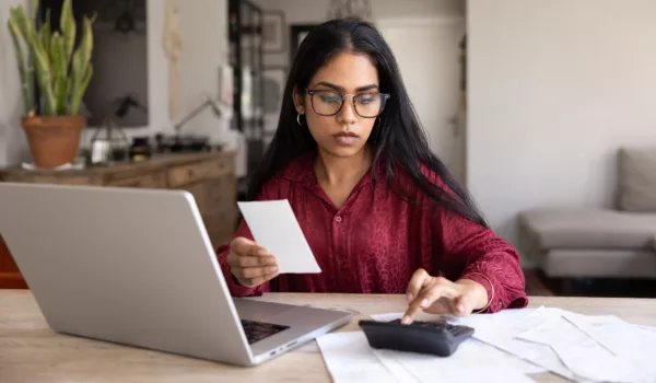Serious young adult Indian girl in eyeglasses calculating budget, checking shopping receipt at laptop at home table, taking control of personal accounting, keeping records of expenses